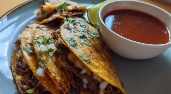 Tre saftiga birria tacos med strimlat kött, lök och koriander, serverade med en skål consommé och en limeklyfta.