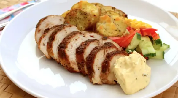 En tallrik med saftig kycklingfilé, skivad och kryddad, serverad med ugnsrostade potatisskivor, majs, paprika och gurka, samt en klick krämig sås.