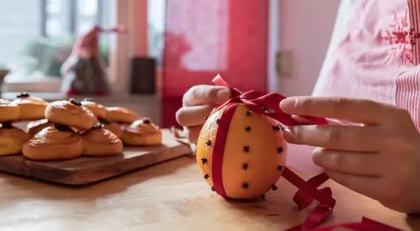 Händer knyter ett rött band runt en apelsin med nejlika hel. Bredvid ligger saffransbullar.