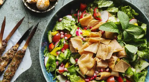 En fräsch fattoush-sallad med krispigt pitabröd, grönsaker och granatäppelkärnor. Bredvid ligger grillspett med kött.