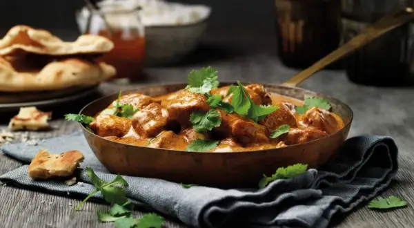 En skål med krämig kyckling tikka masala, toppad med koriander. Serveras med naanbröd och ris.