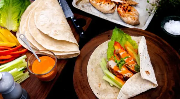 En färdig kyckling wrap med sallad, paprika och gurka. Bredvid ligger tortillabröd, dressing och grillad kyckling.