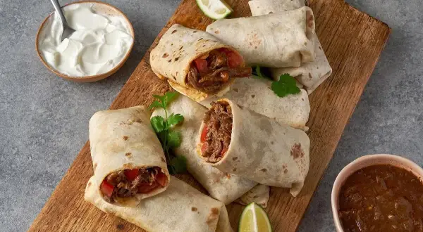 En hög med saftiga chicken burritos på en träskärbräda, serverade med gräddfil, salsa och limeklyftor.