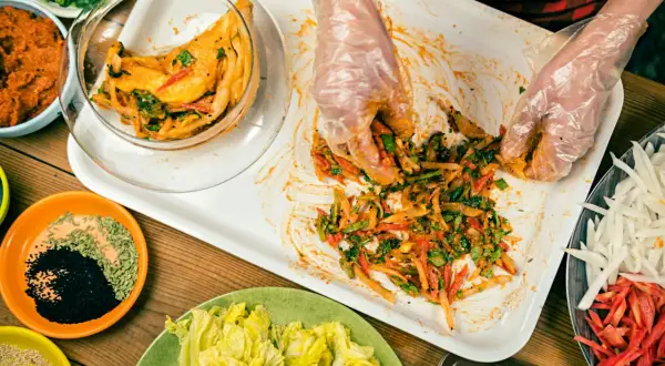 Händer i plasthandskar blandar ingredienser för ett kimchi recept. Grönsaker och kryddor ligger upplagda runt om.