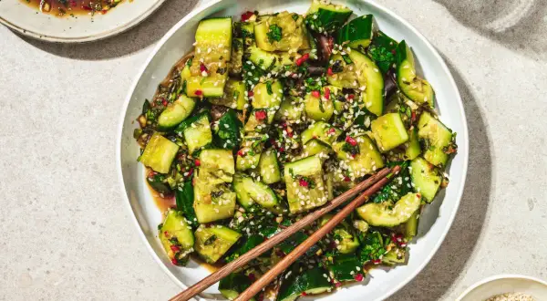 En skål med smashad gurksallad, toppad med sesamfrön och örter, med ätpinnar vilande ovanpå.