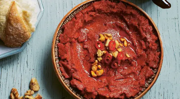 En skål med röd muhammara, toppad med chili och nötter, bredvid en bit bröd. Perfekt för ditt muhammara recept.