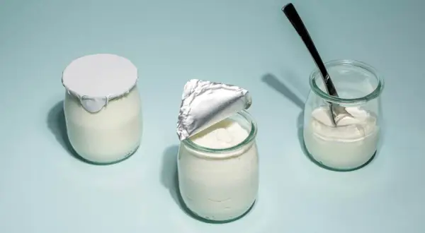 Tre burkar med hemmagjord yoghurt på ljusblå bakgrund. En är helt försluten, en annan är delvis öppnad och en tredje har en sked i sig.