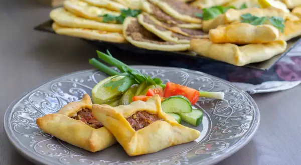 Två lahm bi ajin, små köttfyllda bröd, serveras på en grå tallrik med grönsaker. I bakgrunden syns fler lahm bi ajin och andra bakverk.