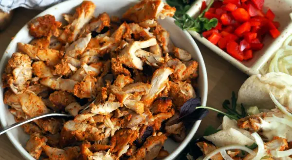 En skål med kryddad kyckling för kyckling gyros, bredvid grönsaker och pitabröd.
