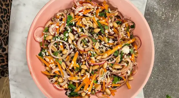 En färgstark ris sallad med jordnötter, morötter, rödlök och örter i en rosa skål.