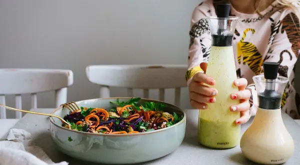 En skål med färgglad sallad och två flaskor dressing, en grön och en ljusgul. En flicka håller i den gröna flaskan.