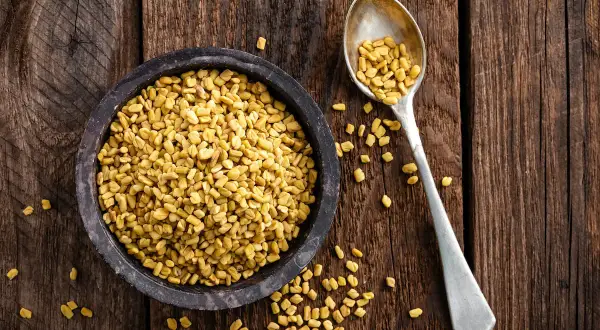 En skål med gyllene bockhornsklöverfrön (fenugreek seeds svenska) bredvid en sked full med samma frön, på en rustik träbakgrund.