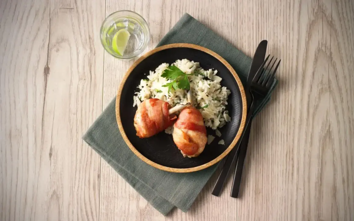 En tallrik med kycklingklubbor inslagna i bacon och krämig risotto. Ett glas vatten med lime och bestick ligger bredvid. Ett perfekt risotto recept kyckling!