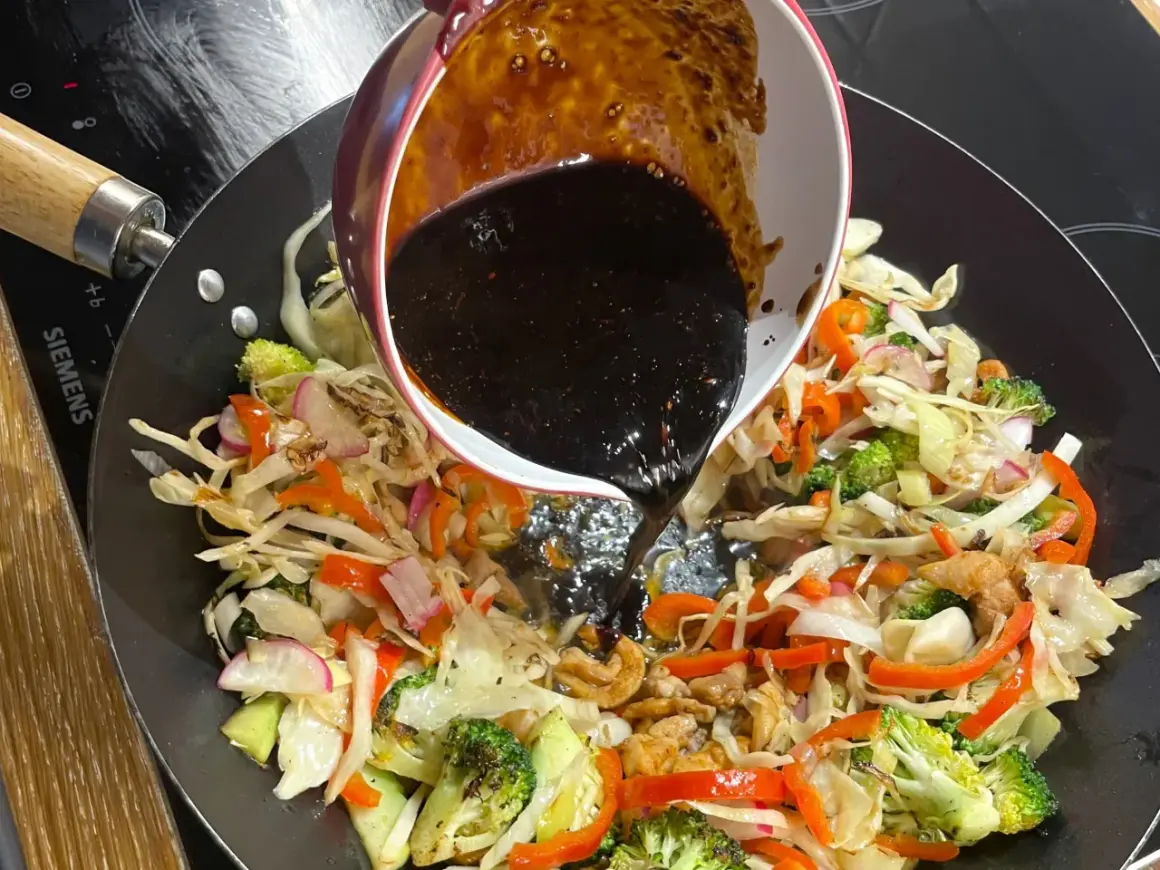 Mörk, glansig wok sås hälls över färgglada grönsaker som broccoli, paprika och kål i en het wokpanna.