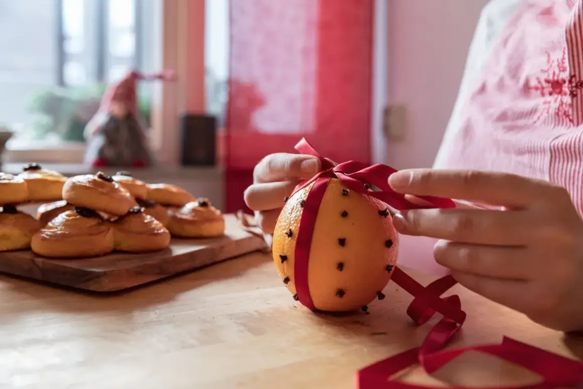 Händer knyter ett rött band runt en apelsin med nejlika hel. Bredvid ligger saffransbullar.