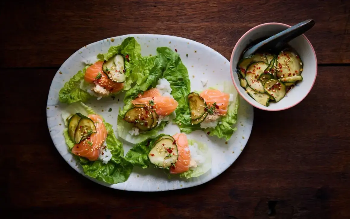 Läckra asiatiska tapas med lax, ris och gurka i salladsblad. En skål med marinerad gurka står bredvid.