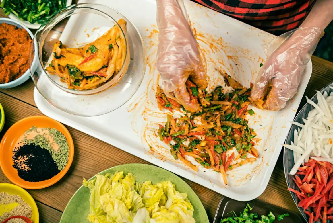 Händer i plasthandskar blandar ingredienser för ett kimchi recept. Grönsaker och kryddor ligger upplagda runt om.