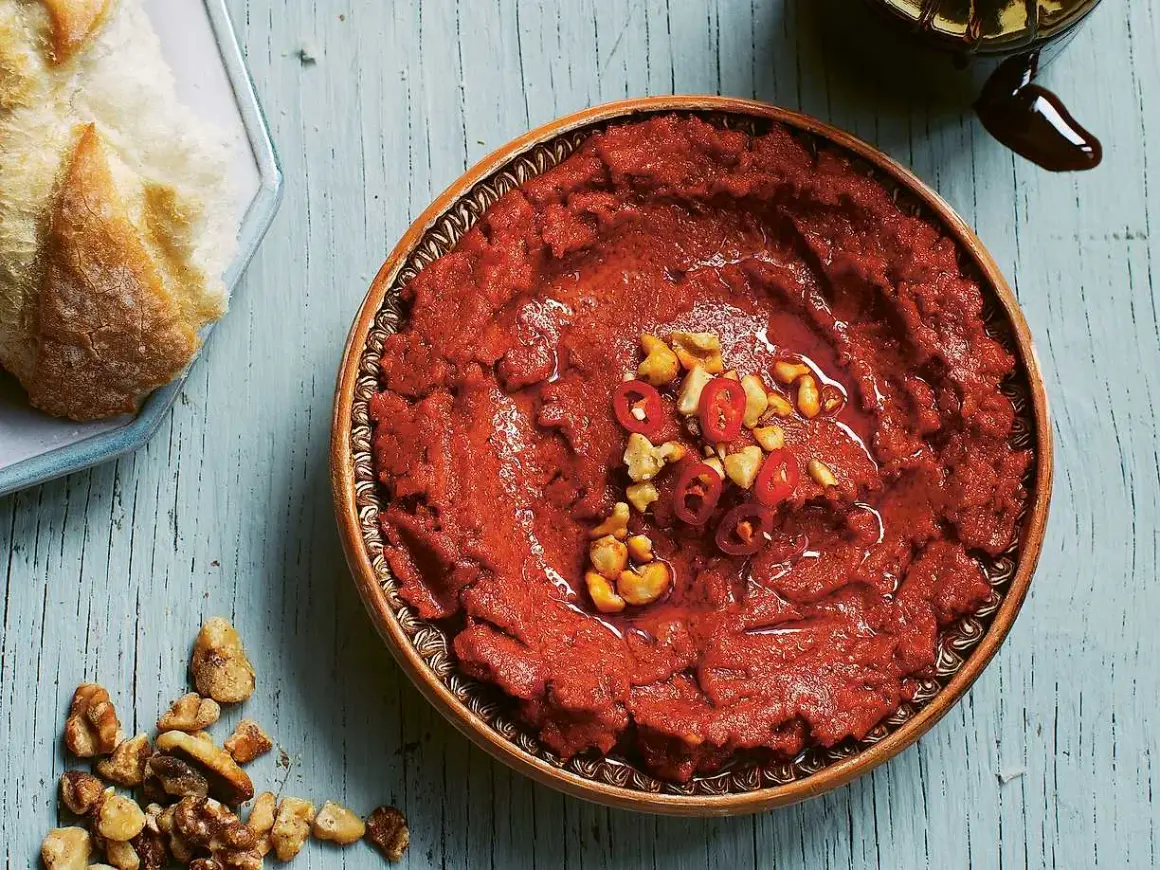 En skål med röd muhammara, toppad med chili och nötter, bredvid en bit bröd. Perfekt för ditt muhammara recept.