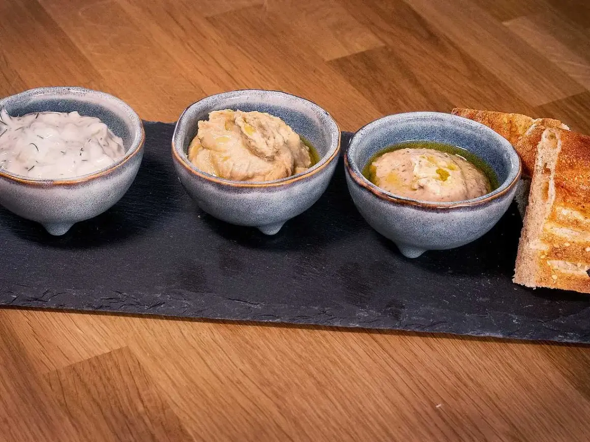 Tre skålar med dippar, som tzatziki och hummus, serveras med bröd. Perfekt för ett meze recept.
