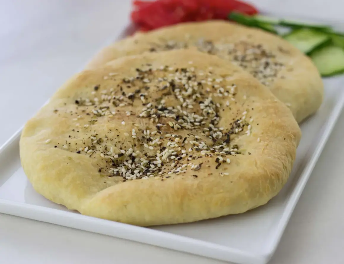 Två runda manakish zaatar med sesamfrön och örter på en vit bricka, med skivad gurka och tomat i bakgrunden.