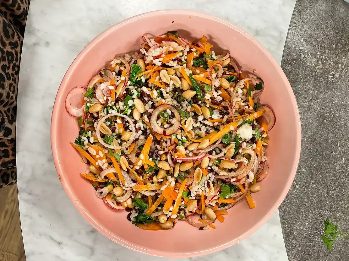 En färgstark ris sallad med jordnötter, morötter, rödlök och örter i en rosa skål.