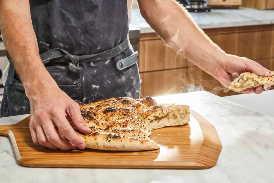 En person i förkläde håller upp en bit av en nybakad, ångande ramazan pidesi. Brödet är gyllene med sesamfrön.