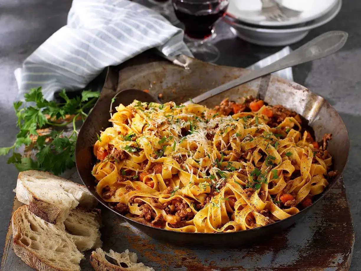 En stor skål med pasta bolognese, toppad med parmesan och persilja, serveras med rustikt bröd och ett glas rödvin.