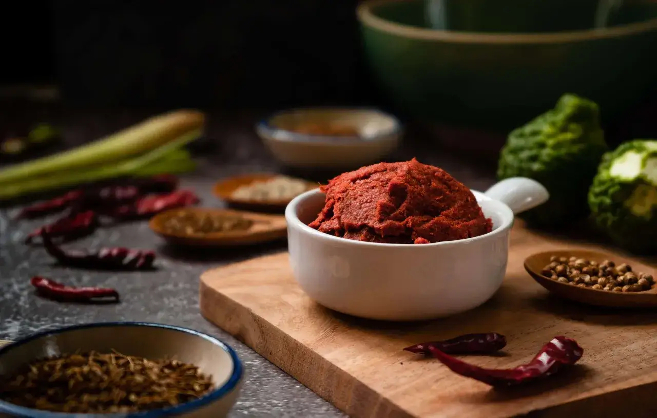 En skål med röd currypasta omgiven av torkade chilifrukter, citrongräs, kaffir lime och kryddor. Ingredienser för en smakrik currypasta.