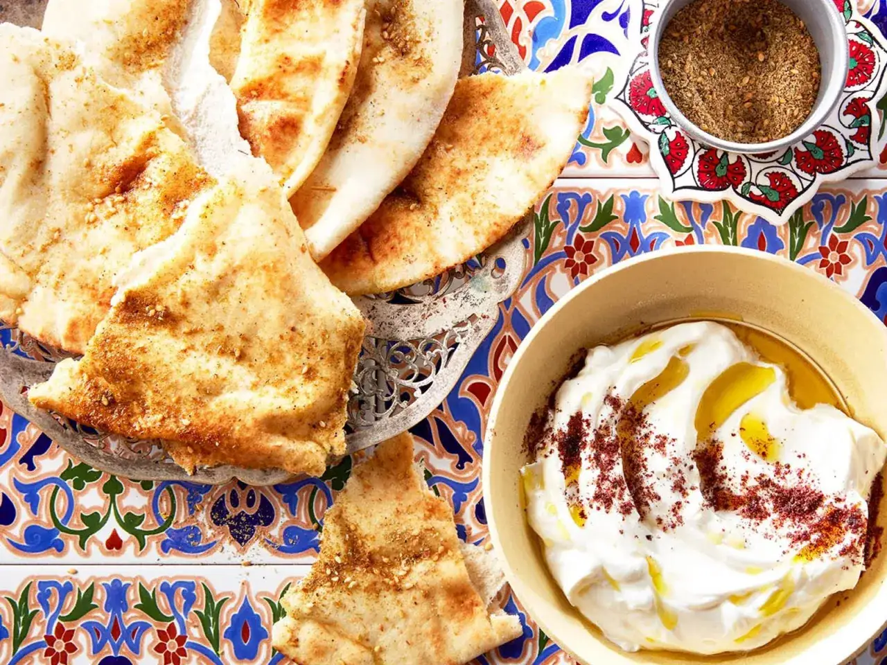 Manakish zaatar, gyllene bröd med kryddig topping, serveras med krämig yoghurt och olivolja på en vackert mönstrad bricka.