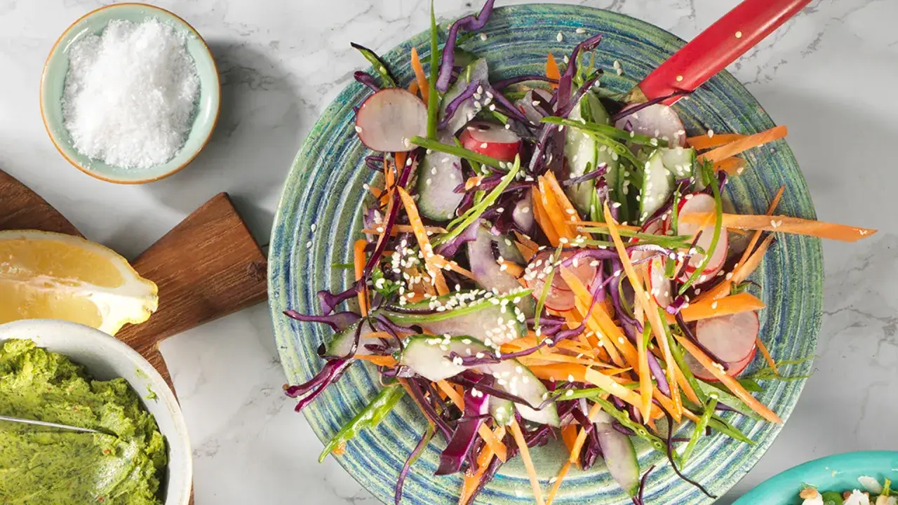 En färgglad asiatisk kålsallad med morötter, rädisor och gurka, toppad med sesamfrön. Serveras med en grön dipp och citron.