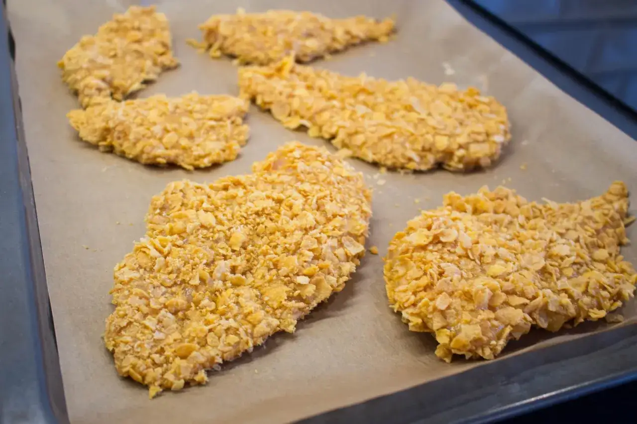 Flera bitar cornflakes kyckling ligger på ett bakplåtspapper, redo att tillagas.