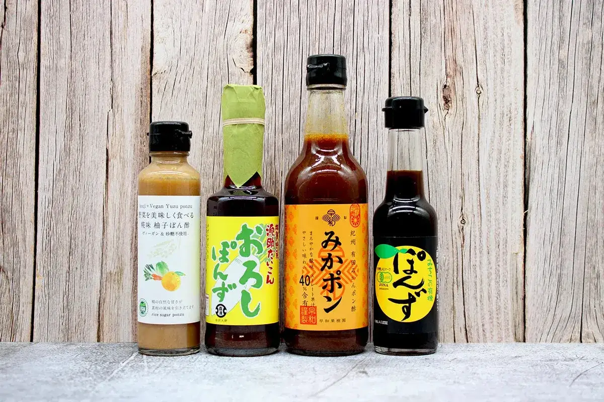 Fyra flaskor med japansk ponzu sås, inklusive en vegansk yuzu-variant, står uppradade mot en träbakgrund.