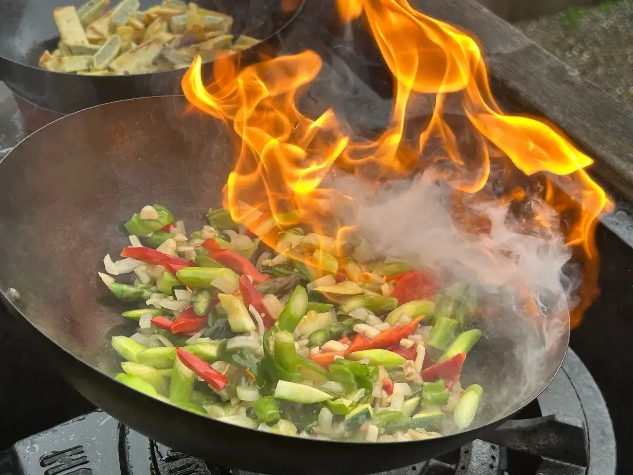 Gr&ouml;nsaker flammande i en wok, med den karakt&auml;ristiska wok hei-doften som sprider sig. I bakgrunden syns mer mat i en annan wok.
