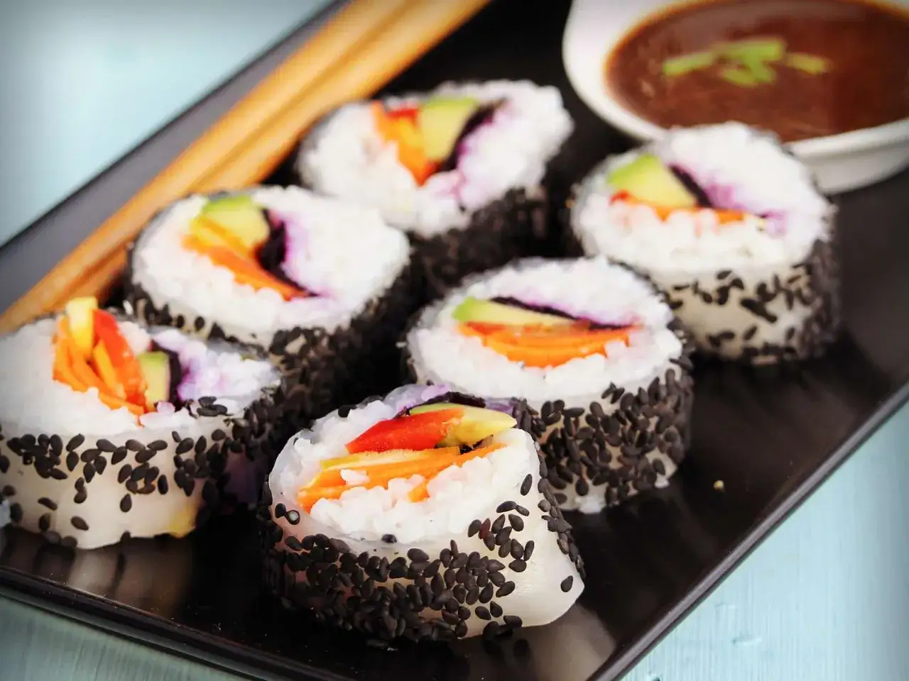 Sju bitar vegansk sushi med avokado och morot, rullade i svarta sesamfr&ouml;n, serveras p&aring; en svart tallrik med en sk&aring;l dipps&aring;s.