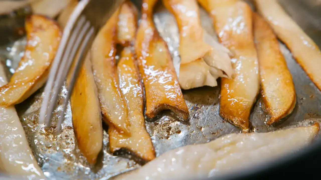 Skivade kungsmusslingar steks gyllene i en panna, redo f&ouml;r ett l&auml;ckert kungsmussling recept. En gaffel syns i bild.