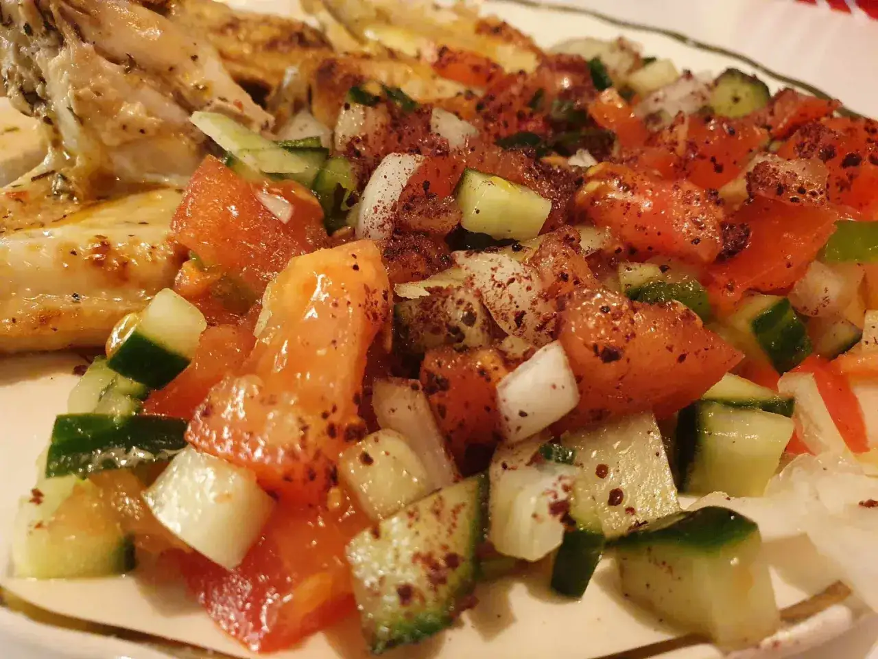 En närbild på en läcker coban salata med saftiga tomater, krispig gurka och lök, kryddad med sumak. Bredvid syns grillad kyckling.