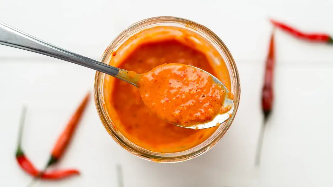 En sked med kryddig piri piri-s&aring;s lyfts ur en burk. Runt burken syns r&ouml;da chilifrukter.