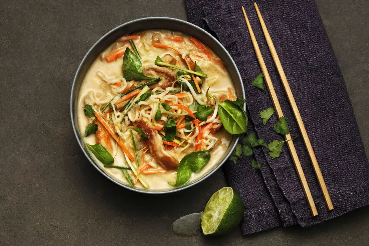 En skål med krämig tom kha gai med kyckling, nudlar, morötter och gröna blad. Ätpinnar och lime vid sidan.