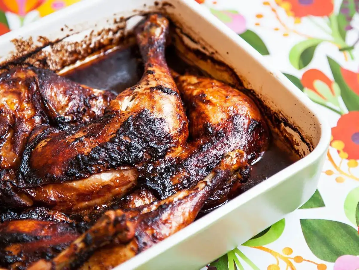Ugnsbakade kycklingben i en mörk, glansig marinad till kycklingben. Serveras i en vit ugnsform på ett blommigt underlag.