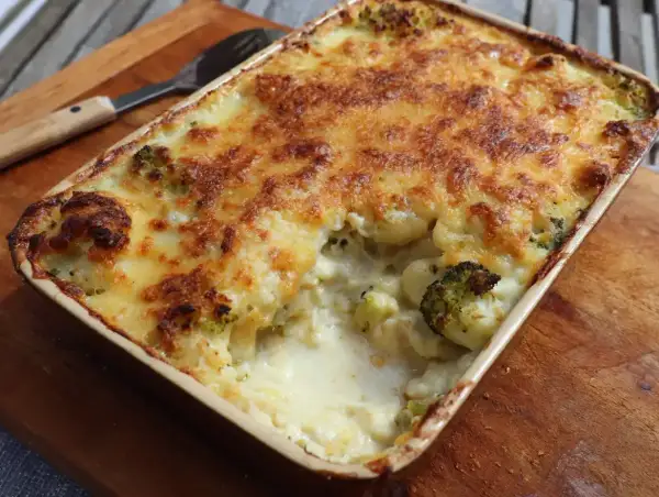 En ugnsform med ostgratinerad blomkål och broccoli, gyllene och bubblande. En sked ligger bredvid.