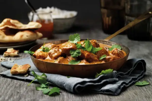 En skål med krämig kyckling tikka masala, toppad med koriander. Serveras med naanbröd och ris.