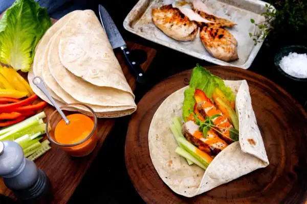 En färdig kyckling wrap med sallad, paprika och gurka. Bredvid ligger tortillabröd, dressing och grillad kyckling.