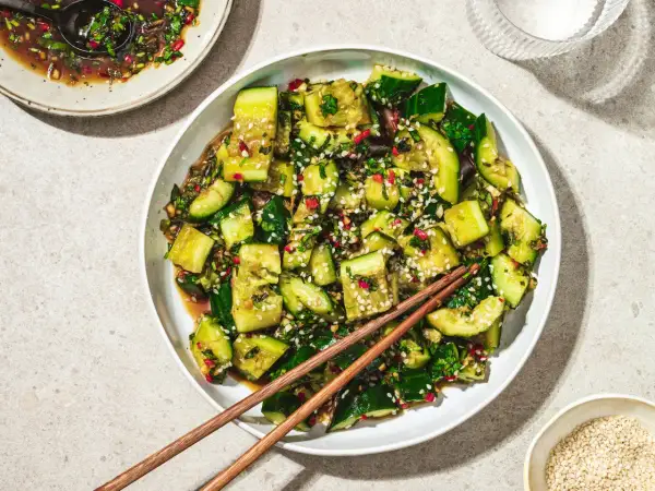En skål med smashad gurksallad, toppad med sesamfrön och örter, med ätpinnar vilande ovanpå.