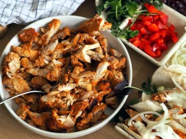 En skål med kryddad kyckling för kyckling gyros, bredvid grönsaker och pitabröd.